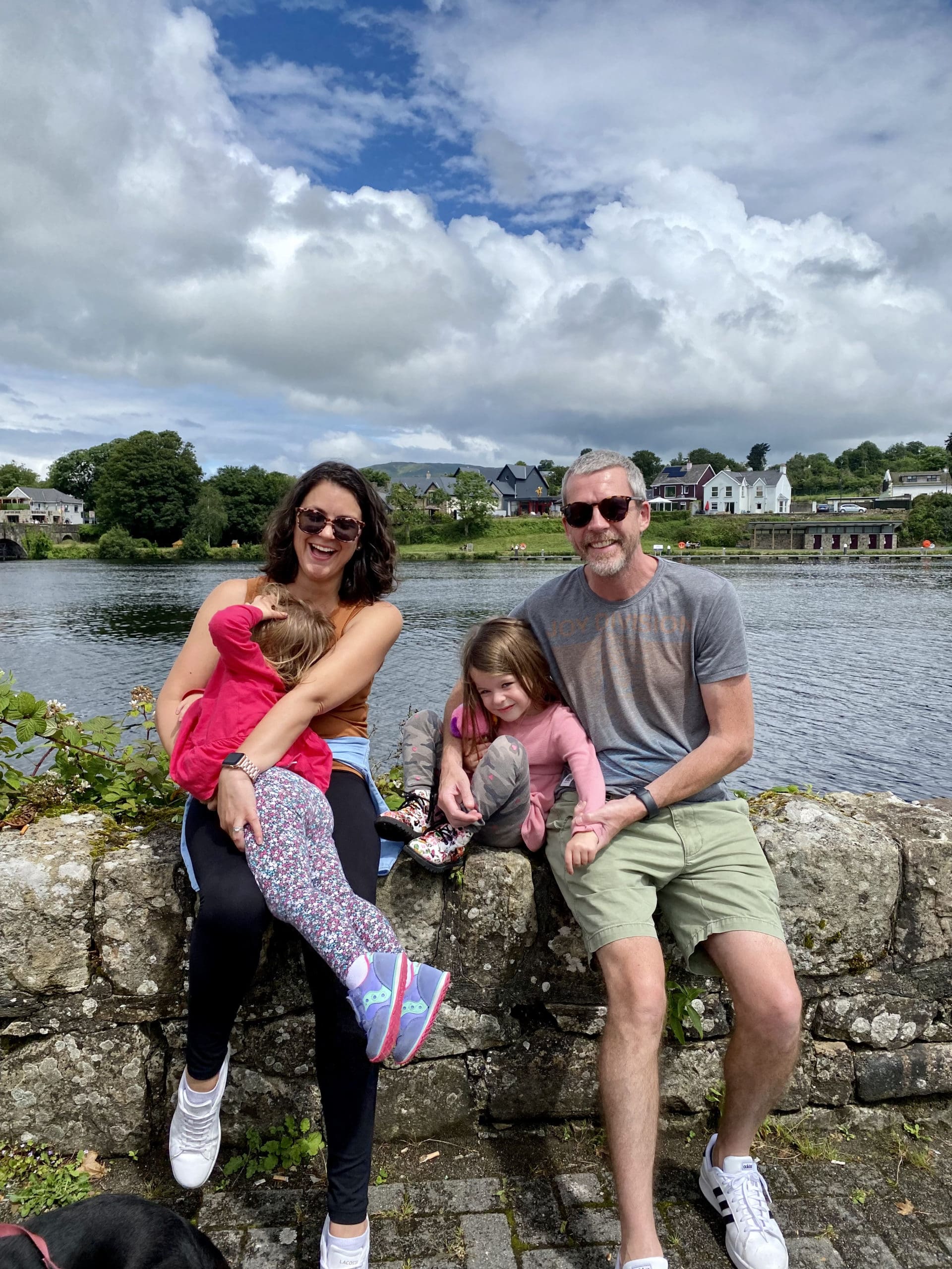 Family sitting on a stone wall by a lakeside on a cloudy day.