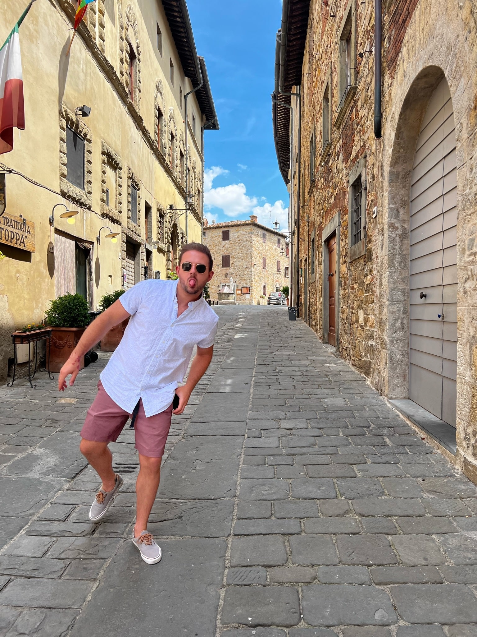 Brennan posing playfully on a cobblestone street in a historic Italian village with stone buildings and blue sky.